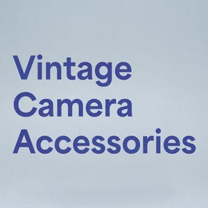 Può includere: Testo blu scuro su sfondo azzurro chiaro: "Vintage Camera Accessories". Le parole sono impilate verticalmente, con "Vintage" in alto, "Camera" al centro e "Accessories" in basso, il tutto in grassetto.