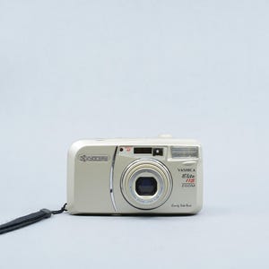 May include: A silver Yashica Elite 115 Zoom camera with a black wrist strap. The camera has a lens, flash, and various buttons. The text "Yashica Elite 115 Zoom" is printed on the front.
