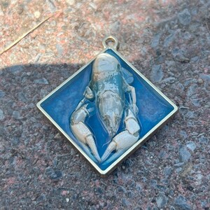 Könnte beinhalten: Goldfarbener Anhänger mit detailliertem Krebs-Design. Der Krebs befindet sich vor einem blauen Hintergrund in einem quadratischen Rahmen. Der Anhänger hat eine Schlaufe für eine Kette.