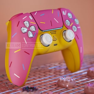 Könnte beinhalten: Ein gelber und rosa PlayStation 5 DualSense-Controller mit einem Streuselmuster. Der Controller befindet sich auf einem Drahtgitter mit rosafarbenen Donuts.