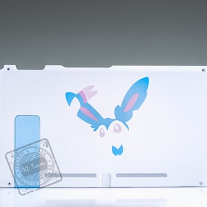 Könnte beinhalten: Weiße Rückplatte für die Nintendo Switch Konsole mit einem blauen und rosa Cartoon-Charakterdesign. Der Charakter hat große Ohren und ein weißes Gesicht mit zwei schwarzen Augen.