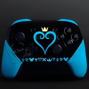 Könnte beinhalten: Ein blauer und schwarzer Videospiel-Controller mit einem Kingdom Hearts-Design. Der Controller verfügt über ein schwarzes Herz mit einer Krone und einem blauen Wirbel sowie eine Reihe von Kingdom Hearts-Symbolen.