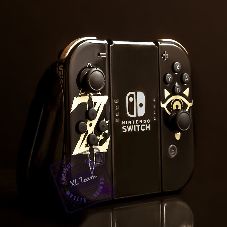 May include: A black Nintendo Switch console with gold accents and a Zelda: Breath of the Wild design. The console has a gold "Z" and a sword design on the left side and a gold eye design on the right side. The console also has the Nintendo Switch logo and text.