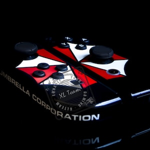 May include: A black Nintendo Switch controller with a custom design featuring the Umbrella Corporation logo, red and white accents, and the text "XL Team".