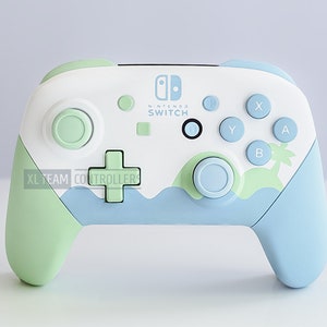May include: A white Nintendo Switch controller with a light blue and green design. The controller has a palm tree and waves on the front. The text "XL TEAM CONTROLLERS" is printed on the front of the controller.