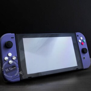 Puede incluir: Una consola Nintendo Switch morada con una pantalla blanca y botones morados y blancos. La consola est&aacute; sobre una superficie de madera oscura.