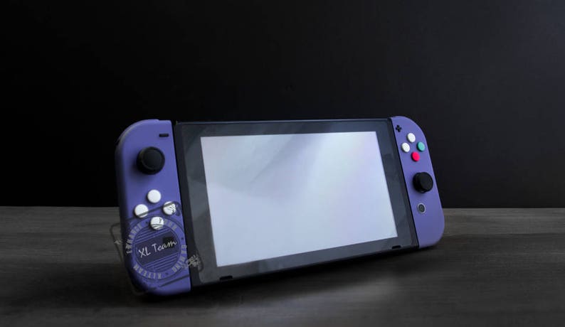 Puede incluir: Una consola Nintendo Switch morada con una pantalla blanca y botones morados y blancos. La consola est&aacute; sobre una superficie de madera oscura.