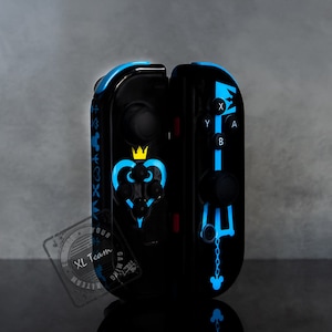 Op de afbeelding: Zwarte en blauwe gamecontroller met een hart- en kroonontwerp. De controller heeft blauwe accenten en symbolen, waaronder een sleutelvormig ontwerp. De knoppen zijn gelabeld met witte letters. Voor een gameconsole.