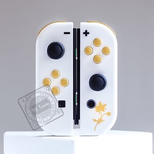 K&ouml;nnte beinhalten: Ein wei&szlig;er Nintendo Switch Joy-Con-Controller mit goldenen Kn&ouml;pfen und einem floralen Design. Der Controller hat einen schwarzen Knopf links und einen schwarzen Knopf rechts.