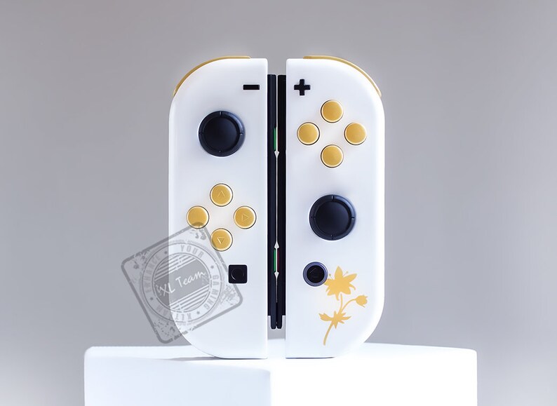 K&ouml;nnte beinhalten: Ein wei&szlig;er Nintendo Switch Joy-Con-Controller mit goldenen Kn&ouml;pfen und einem floralen Design. Der Controller hat einen schwarzen Knopf links und einen schwarzen Knopf rechts.