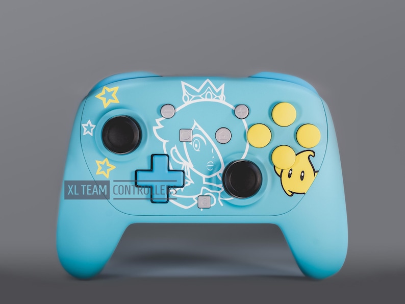 May include: A light blue video game controller featuring a white line art illustration of a character with a crown, yellow star graphics, and a yellow Luma character. The controller includes black joysticks, yellow buttons, and the text XL TEAM CONTROLLERS.