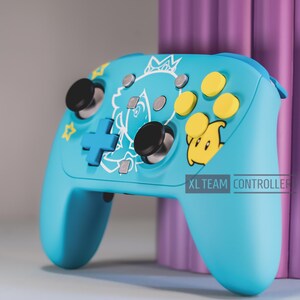 May include: A light blue wireless video game controller featuring a white line art graphic of a character wearing a crown, yellow action buttons, a yellow star icon, black joysticks, and a blue directional pad. The text XL TEAM CONTROLLER is visible.