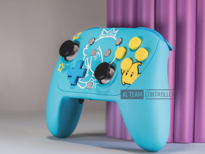 May include: A light blue wireless video game controller featuring a white line art graphic of a character wearing a crown, yellow action buttons, a yellow star icon, black joysticks, and a blue directional pad. The text XL TEAM CONTROLLER is visible.