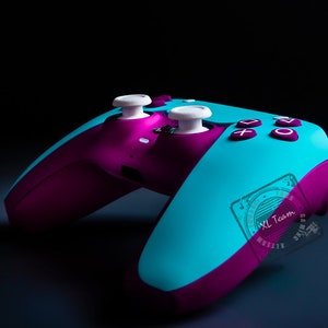 Può includere: Un controller per videogiochi color turchese e viola con pulsanti bianchi. Il controller ha un adesivo sul lato che dice "XL Team".