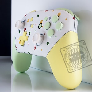 May include: A white and yellow custom-designed video game controller with a green and white speckled pattern. The controller has a yellow button and a green button. The controller is on a white surface.