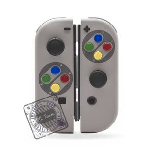 May include: A gray Nintendo Switch console with a custom Super Nintendo-style design. The console has a black and white color scheme with colorful buttons. The console has a black and white sticker on the bottom that says "Enhance Your Gaming XL Team".