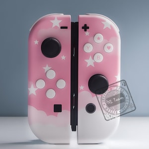 Könnte beinhalten: Ein rosa-weißer Nintendo Switch-Controller mit einem Wolken- und Sternendesign. Der Controller hat auf jeder Seite einen schwarzen Knopf und einen schwarzen Joystick.