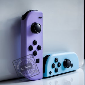 Könnte beinhalten: Zwei Nintendo Switch-Controller, einer lila und einer hellblau. Die Controller haben schwarze Knöpfe und Joysticks.