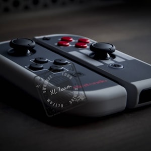 Może przedstawiać: Szary i czarny kontroler Nintendo Switch z szaro-białą płytką czołową. Płytka czołowa ma nadrukowany tekst "Enhance Your Gaming" i "XL Team". Kontroler ma czarny joystick i cztery czerwone przyciski.