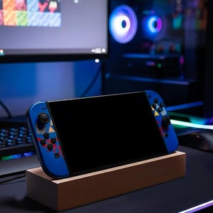 Puede incluir: Consola de juegos port&aacute;til azul con pantalla negra, apoyada en un soporte de madera. La consola tiene mandos azules con detalles coloridos. Un teclado, un rat&oacute;n y un monitor est&aacute;n en el fondo, lo que sugiere una configuraci&oacute;n de juego.
