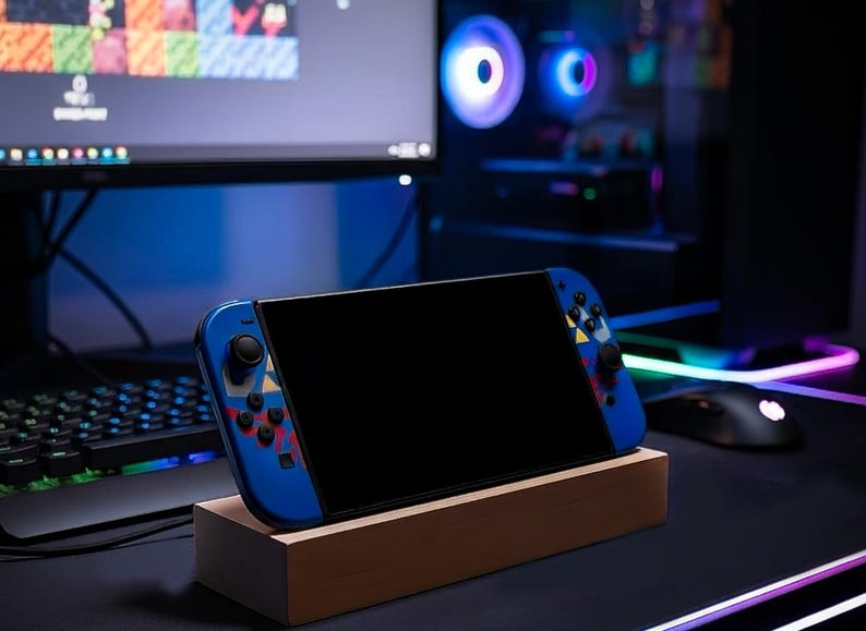 Puede incluir: Consola de juegos port&aacute;til azul con pantalla negra, apoyada en un soporte de madera. La consola tiene mandos azules con detalles coloridos. Un teclado, un rat&oacute;n y un monitor est&aacute;n en el fondo, lo que sugiere una configuraci&oacute;n de juego.