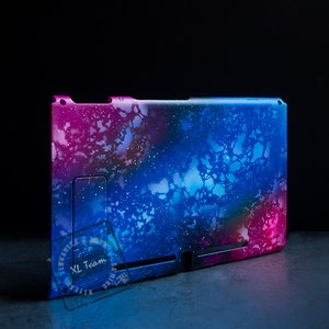 May include: A tablet case with a vibrant galaxy design in shades of blue, pink, and purple. The case features a stand and a cutout for the tablet's features. The XL Team logo is visible on the lower left corner.