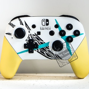 Peut inclure: Une manette Nintendo Switch blanche avec un design géométrique noir et turquoise. La manette a des poignées jaunes et le logo Nintendo Switch est visible sur le devant.