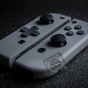 Könnte beinhalten: Ein Paar graue Gamecontroller mit schwarzen Knöpfen und Joysticks. Die Controller haben ein kreuzförmiges Steuerkreuz und Aktionstasten mit Symbolen. Die Controller sind für den Anschluss an eine Konsole konzipiert.