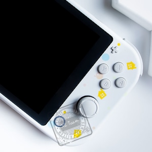 Może przedstawiać: Biała przenośna konsola do gier z czarnym ekranem i kolorowymi przyciskami. Konsola ma biało-żółty wzór z małymi ikonami domów.