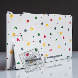 May include: White Nintendo Switch console with a custom design featuring a repeating pattern of green leaves, yellow houses, red apples, and gray circles. The design is inspired by the popular video game Animal Crossing.