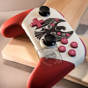 May include: A white and red Nintendo Switch controller with a black eagle design and pink buttons. The controller has a red and white design with a black eagle on the front. The buttons are pink and the controller is on a wooden surface.