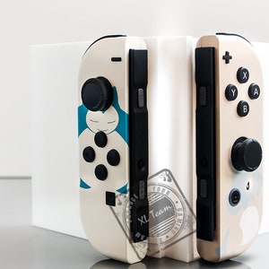 May include: Two white Nintendo Switch Joy-Con controllers with custom designs. The left controller features a blue and white character with a sleepy face. The right controller has a beige and white design with a black button.