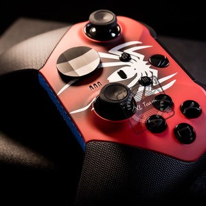May include: A red and black Xbox controller with a spider design. The controller has a textured grip and a black faceplate.