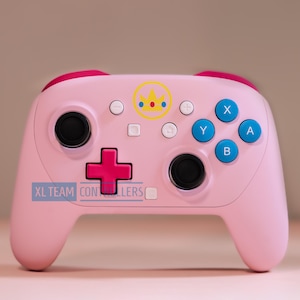 May include: A light pink wireless video game controller featuring a yellow crown icon with red and blue accents at the top center. The device includes a dark pink directional pad, two black joysticks, and four blue action buttons labeled A, B, X, and Y.