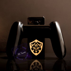 May include: Black Nintendo Switch console stand with two black Joy-Con controllers and a gold Triforce shield design. The stand is labeled "XL Team Gaming" in a purple circle.