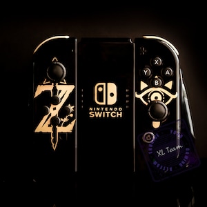 May include: A black Nintendo Switch console with gold accents. The console has a custom design featuring the "Z" symbol from the Legend of Zelda video game series. The console also has a custom design on the right joy-con featuring the "Triforce" symbol from the Legend of Zelda video game series. The console is on a black surface.