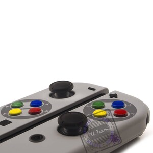 May include: A gray and white video game controller with black buttons and a red, yellow, blue, and green button layout. The controller is split in half and has a black joystick on each side.