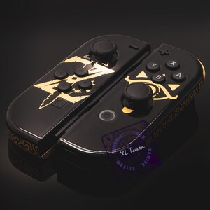 May include: Black Nintendo Switch Joy-Con controllers with gold accents and a Zelda design. The left Joy-Con features a stylized image of the Master Sword, while the right Joy-Con features the Triforce symbol and the Eye of Ganondorf.