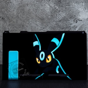 Könnte beinhalten: Schwarze Spielekonsole mit einem blauen und schwarzen Pokemon-Charakter-Grafik auf dem Bildschirm. Die Konsole hat ein blaues Licht auf der linken Seite.