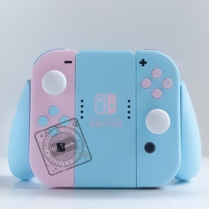 Könnte beinhalten: Eine hellblaue und rosafarbene Nintendo Switch Konsole mit Joy-Cons. Die Konsole hat das Nintendo Switch Logo und den Text "Nintendo Switch" aufgedruckt. Die Joy-Cons haben weiße und rosafarbene Knöpfe.