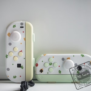 May include: Two white and green Nintendo Switch Joy-Con controllers with a  "#XL Team" watermark. The controllers feature a  "Animal Crossing: New Horizons" design with a variety of  icons and patterns.