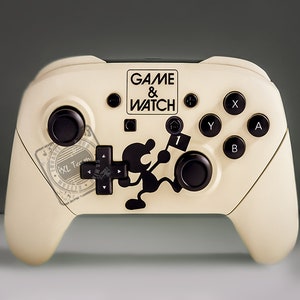 May include: Cream-colored video game controller with black buttons and a black Game & Watch graphic. The controller has the text "GAME & WATCH" and a black character holding a flag. The controller also has a black directional pad.