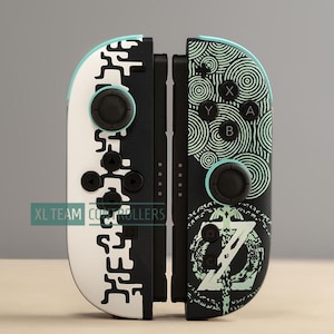 May include: Two game controllers featuring a black and white color scheme. The left controller has a white base with black abstract patterns. The right controller is black with green circular line art and a sword emblem. Text reads XL TEAM CONTROLLERS.