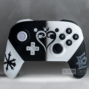 May include: A black and white custom Nintendo Switch controller with a Kingdom Hearts design. The controller features a black and white heart with a crown on top, a black and white symbol on the left side, and a black and white symbol on the right side. The text "XL TEAM CONTROLLERS" is printed on the bottom of the controller.