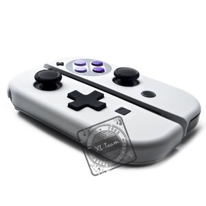 May include: A white Nintendo Switch controller with a Super Nintendo Entertainment System (SNES) design. The controller has a black d-pad, black buttons, and a gray faceplate with four purple buttons. The controller has a black XL Team logo on the bottom.