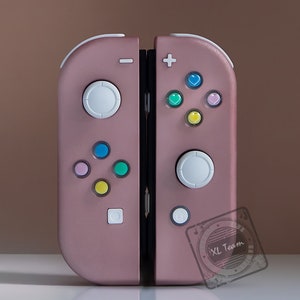 Op de afbeelding: Een roze gouden Nintendo Switch-controller met witte knoppen en kleurrijke hartvormige knoppen. De controller heeft een zwarte naad in het midden en de tekst "XL Team" is zichtbaar in de rechterbenedenhoek.