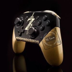 Könnte beinhalten: Ein schwarzer und goldener Nintendo Switch-Controller mit einem goldenen Zelda-Design und einem goldenen "XL Team"-Stempel auf der rechten Seite.