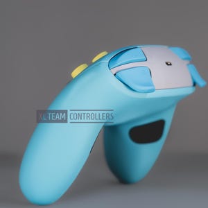 May include: A light blue video game controller featuring a silver top panel, light blue shoulder buttons, and yellow top buttons. The text XL TEAM CONTROLLERS is printed in a rectangular box on the side of the device handle.