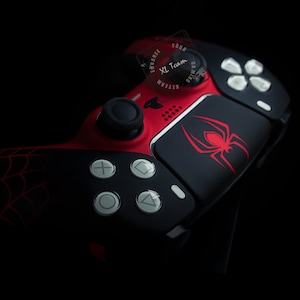 May include: A black and red PlayStation controller with a spider web design and a red spider logo on the touchpad. The controller has a black and white button layout.
