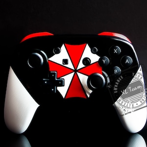May include: A black and white Nintendo Switch controller with a red and white Umbrella Corporation logo. The controller has white grips and black buttons. The logo is a red umbrella with a white background and a black border.